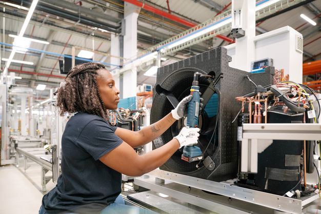 Eine Person montiert ein Bauteil an einer Wärmepumpe in einer Fabrikhalle. 